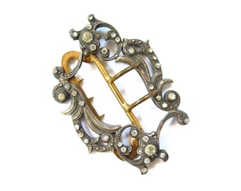 May include: An antique silver-tone belt buckle with a gold-tone center bar. The buckle is adorned with clear, round gemstones and features an ornate, floral design. The buckle is set against a white background.