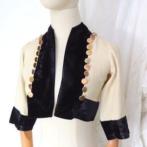 May include: Cream bolero jacket with black velvet trim and collar. The jacket has short sleeves and is decorated with gold-coloured circular buttons. A vintage-style garment. The jacket's measurements are in centimetres.