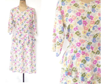 Könnte beinhalten: Ein knielanges Kleid mit weißem Grund und einem mehrfarbigen Blumenmuster in Rosa, Blau, Gelb und Grün. Das Kleidungsstück hat Dreiviertelärmel, einen Rundhalsausschnitt und ein Schößchen an der Taille über einem gerade geschnittenen Rock.