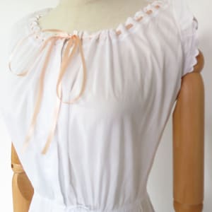 Op de afbeelding: Een witte katoenen chemise met een geplooide halslijn en een strik van perzikkleurig satijnen lint. De chemise heeft korte, gekrulde mouwen en een tailleband.