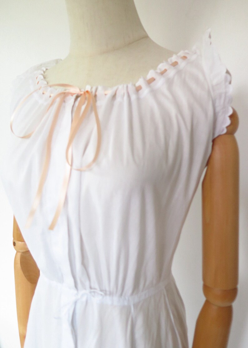 Op de afbeelding: Een witte katoenen chemise met een geplooide halslijn en een strik van perzikkleurig satijnen lint. De chemise heeft korte, gekrulde mouwen en een tailleband.