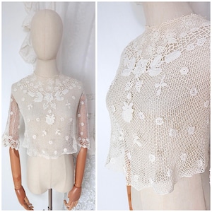 Puede incluir: Una blusa de ganchillo de color blanco roto con mangas cortas y un complejo patrón de malla calada. La prenda está adornada con delicados motivos florales y diseños de hojas integrados en el encaje, con cuello alto y bordes festoneados.
