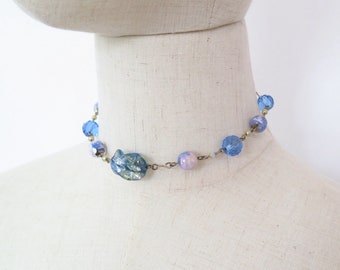 May include: A choker necklace featuring a series of blue and pink beads strung on a thin metal chain. The beads include faceted blue glass, smooth iridescent pink spheres, and a central irregular blue piece with a textured, metallic finish.