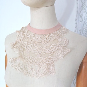 May include: A vintage-style, blush pink lace collar. The collar features multiple layers of delicate, intricately patterned lace, attached to a soft pink satin ribbon. The collar is displayed on a mannequin, showcasing its elegant design.