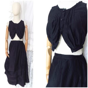 Op de afbeelding: Een tweedelige zwarte outfit bestaande uit een mouwloze, aan de voorzijde geknoopte cropped top met geplooide details bij de halslijn en een bijpassende midi-rok met hoge taille. De set is volledig zwart en voorzien van zichtbare knopen.