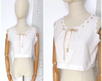 Puede incluir: Top blanco sin mangas con escote festoneado y lazo de cinta de raso. El top tiene una botonadura frontal y se exhibe en un maniquí. El top es de estilo crop y parece estar hecho de algodón.