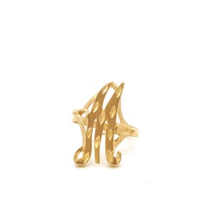 May include: A gold ring featuring a prominent letter M as the centerpiece. The letter M is crafted with a textured, diamond-cut finish that creates a shimmering effect across its surface. The band is thin and connects to the sides of the letter.