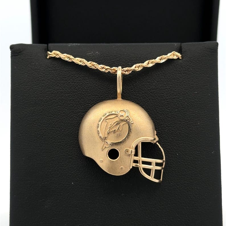 K&ouml;nnte beinhalten: Ein goldfarbener Anh&auml;nger in Form eines Football-Helms mit einem Delfin-Logo an der Seite und einem Gesichtsschutz, der an einer passenden goldfarbenen Kordelkette h&auml;ngt. Der Helm hat eine matte Oberfl&auml;che und liegt auf einem dunklen Untergrund.
