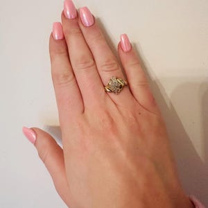 May include: A gold ring with a large, clear, round stone surrounded by smaller stones. The ring is on a hand with pink nail polish.