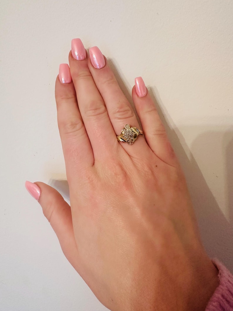 May include: A gold ring with a large, clear, round stone surrounded by smaller stones. The ring is on a hand with pink nail polish.