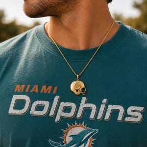 K&ouml;nnte beinhalten: Ein goldfarbener Metallanh&auml;nger in Form eines Footballhelms mit dem Miami Dolphins-Logo an der Seite, der an einer d&uuml;nnen goldfarbenen Kette h&auml;ngt. Der Anh&auml;nger liegt auf einem blaugr&uuml;nen T-Shirt mit dem in Wei&szlig; gedruckten Text MIAMI DOLPHINS.