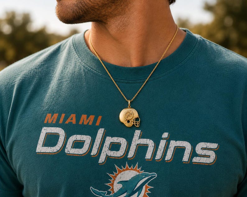 K&ouml;nnte beinhalten: Ein goldfarbener Metallanh&auml;nger in Form eines Footballhelms mit dem Miami Dolphins-Logo an der Seite, der an einer d&uuml;nnen goldfarbenen Kette h&auml;ngt. Der Anh&auml;nger liegt auf einem blaugr&uuml;nen T-Shirt mit dem in Wei&szlig; gedruckten Text MIAMI DOLPHINS.