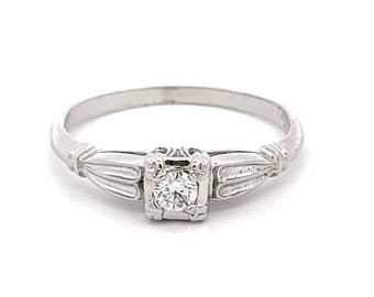 May include: A silver-toned metal ring featuring a single round, clear faceted stone set in a square-shaped mounting. The band displays a decorative, ribbed design on both sides of the central setting, creating a structured and symmetrical appearance.