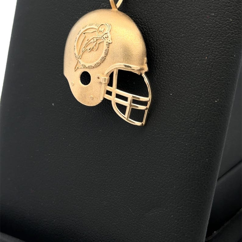 K&ouml;nnte beinhalten: Ein goldfarbener Anh&auml;nger in Form eines Footballhelms mit matter Oberfl&auml;che. Die Seite des Helms zeigt ein eingraviertes Logo eines Delfins, der durch einen sonnenstrahlenf&ouml;rmigen Kreis springt, mit dem Buchstaben M oben. Inklusive detailliertem Gesichtsschutz.