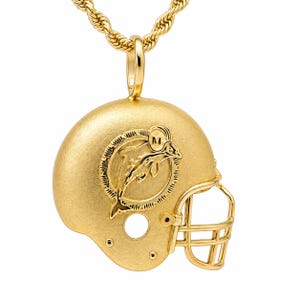 K&ouml;nnte beinhalten: Ein goldfarbener Anh&auml;nger in Form eines Footballhelms, der an einer passenden Kordelkette h&auml;ngt. Der Helm hat eine matt strukturierte Oberfl&auml;che und ein erhabenes Emblem eines springenden Delfins in einem Kreis, mit dem Buchstaben M auf dem Delfin.