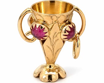 Könnte beinhalten: Ein goldener Kelch mit zwei geschwungenen Henkeln und einem blütenförmigen Fuß. Die Außenseite ist mit gravierten weizenähnlichen Mustern und zwei markanten, runden, dunkelrosa Edelsteinen in gezackten Goldfassungen verziert.