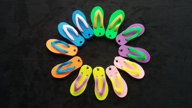 May include: A collection of ten colourful flip-flops arranged in a circle on a black background. The flip-flops are made of rubber and have a variety of colours, including blue, green, orange, pink, and yellow.