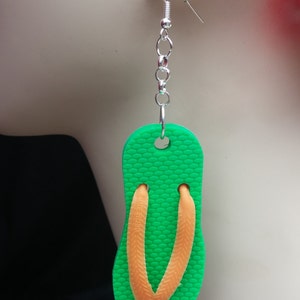 May include: Green flip flop shaped earrings with orange straps. The earrings are hanging from silver chains.