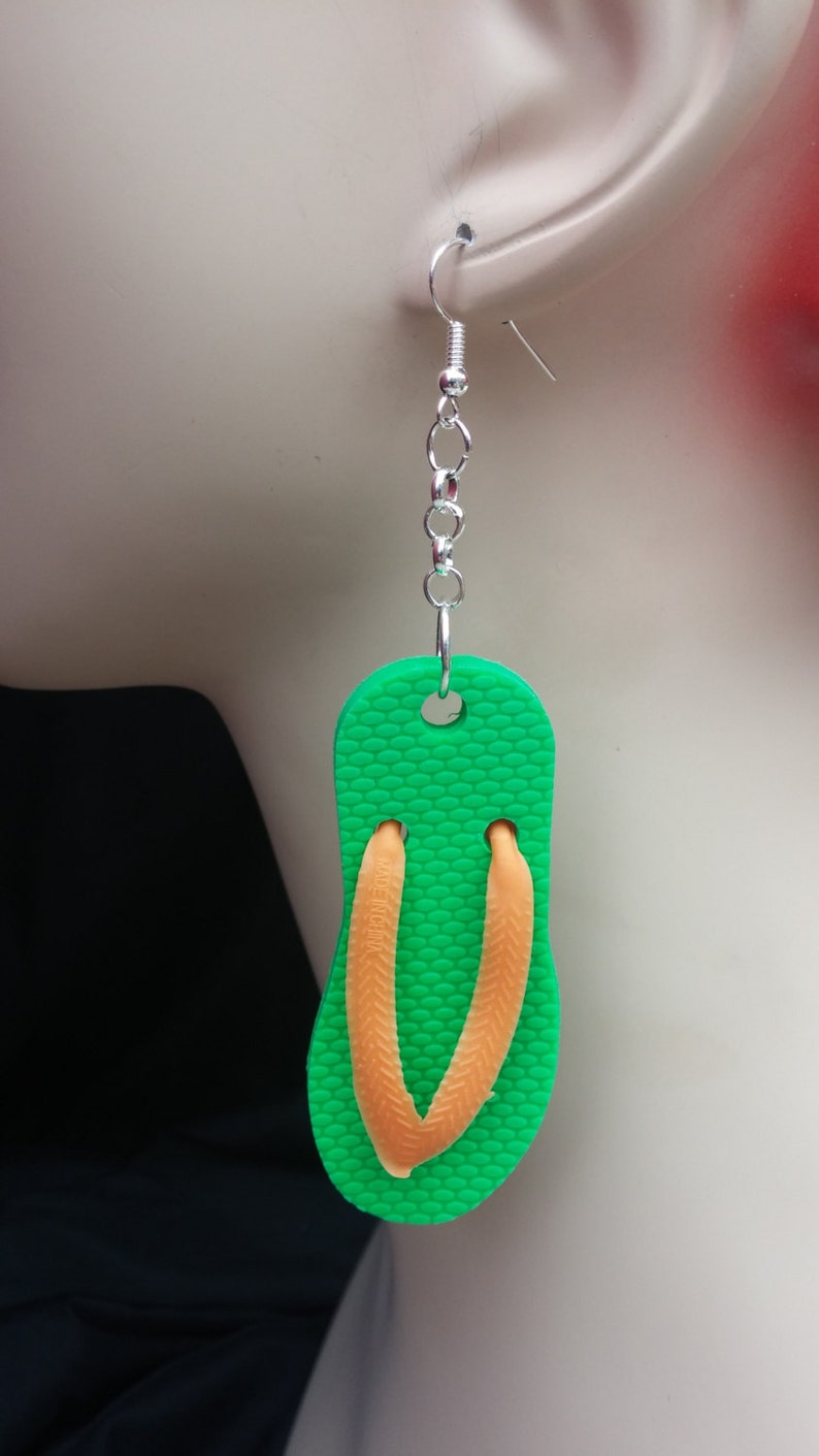 May include: Green flip flop shaped earrings with orange straps. The earrings are hanging from silver chains.