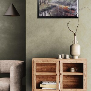 Op de afbeelding: Een ingelijst landschapsschilderij van een mistige bossc&egrave;ne met een rivier. Een houten kast met glazen deuren bevat boeken en een mok. Een vaas met takken en kleine witte kopjes staat erop. Een moderne hanglamp.