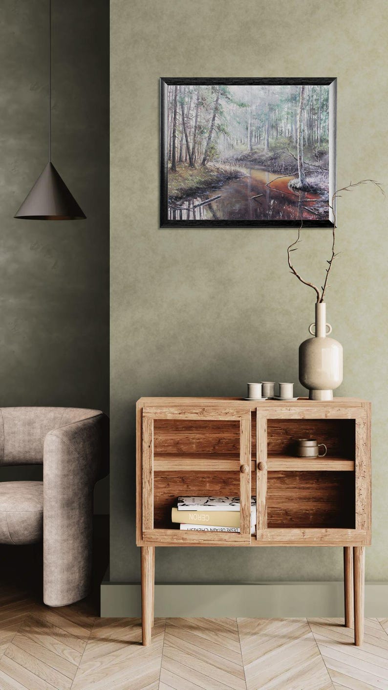 Op de afbeelding: Een ingelijst landschapsschilderij van een mistige bossc&egrave;ne met een rivier. Een houten kast met glazen deuren bevat boeken en een mok. Een vaas met takken en kleine witte kopjes staat erop. Een moderne hanglamp.