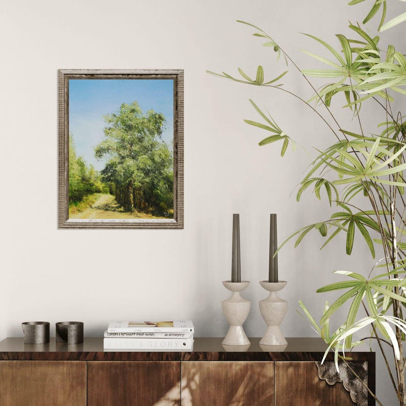 May include: A framed landscape painting depicts a dirt path leading into a forest, with a large tree in the center. The painting has a light blue sky and green foliage. The frame is a light brown color. The painting is displayed on a dark wood cabinet.