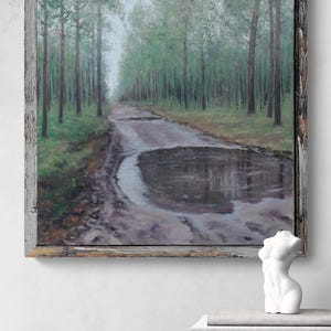 May include: An oil painting depicting a forest path with a large puddle reflecting the sky, framed in a rustic wooden frame. A white torso sculpture and books are on a wooden table below the painting.