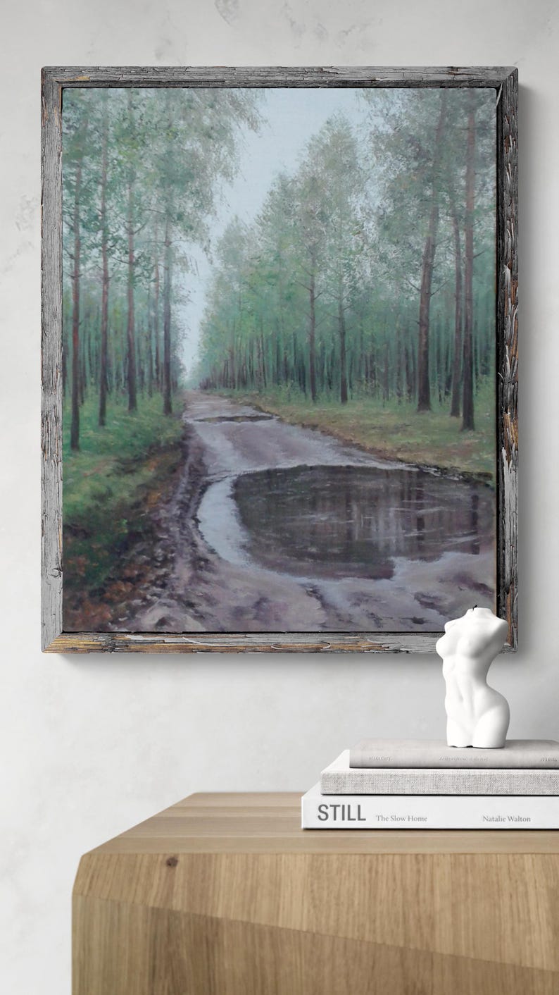 May include: An oil painting depicting a forest path with a large puddle reflecting the sky, framed in a rustic wooden frame. A white torso sculpture and books are on a wooden table below the painting.