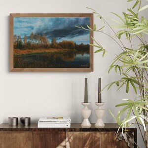 Pu&ograve; includere: Un dipinto di paesaggio incorniciato raffigura una serena scena lacustre sotto un cielo drammatico. L'opera presenta un cielo scuro e tempestoso sopra uno specchio d'acqua riflettente, con alberi che costeggiano la riva lontana. La cornice &egrave; in un caldo tono legno naturale.