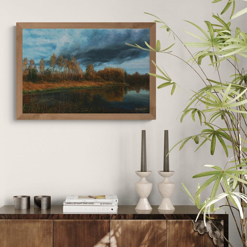 Pu&ograve; includere: Un dipinto di paesaggio incorniciato raffigura una serena scena lacustre sotto un cielo drammatico. L'opera presenta un cielo scuro e tempestoso sopra uno specchio d'acqua riflettente, con alberi che costeggiano la riva lontana. La cornice &egrave; in un caldo tono legno naturale.