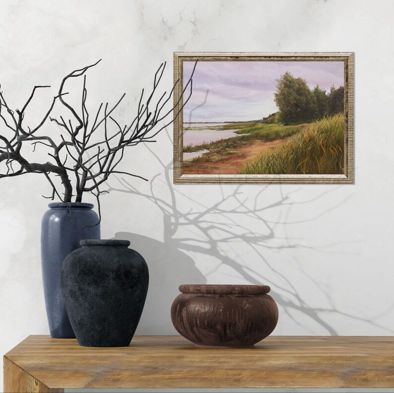 May include: A framed landscape painting shows a waterside scene under a cloudy sky. A tall blue vase, a dark textured vase, and a small brown bowl are on a wooden surface. Black branches are in the blue vase. The painting is in a wooden frame.