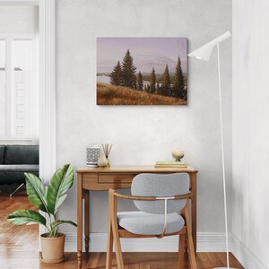 May include: A landscape painting depicts a serene scene with evergreen trees, a body of water, and a golden field under a soft, cloudy sky. The artwork is displayed above a wooden desk with a gray chair, a potted plant, and a modern floor lamp.