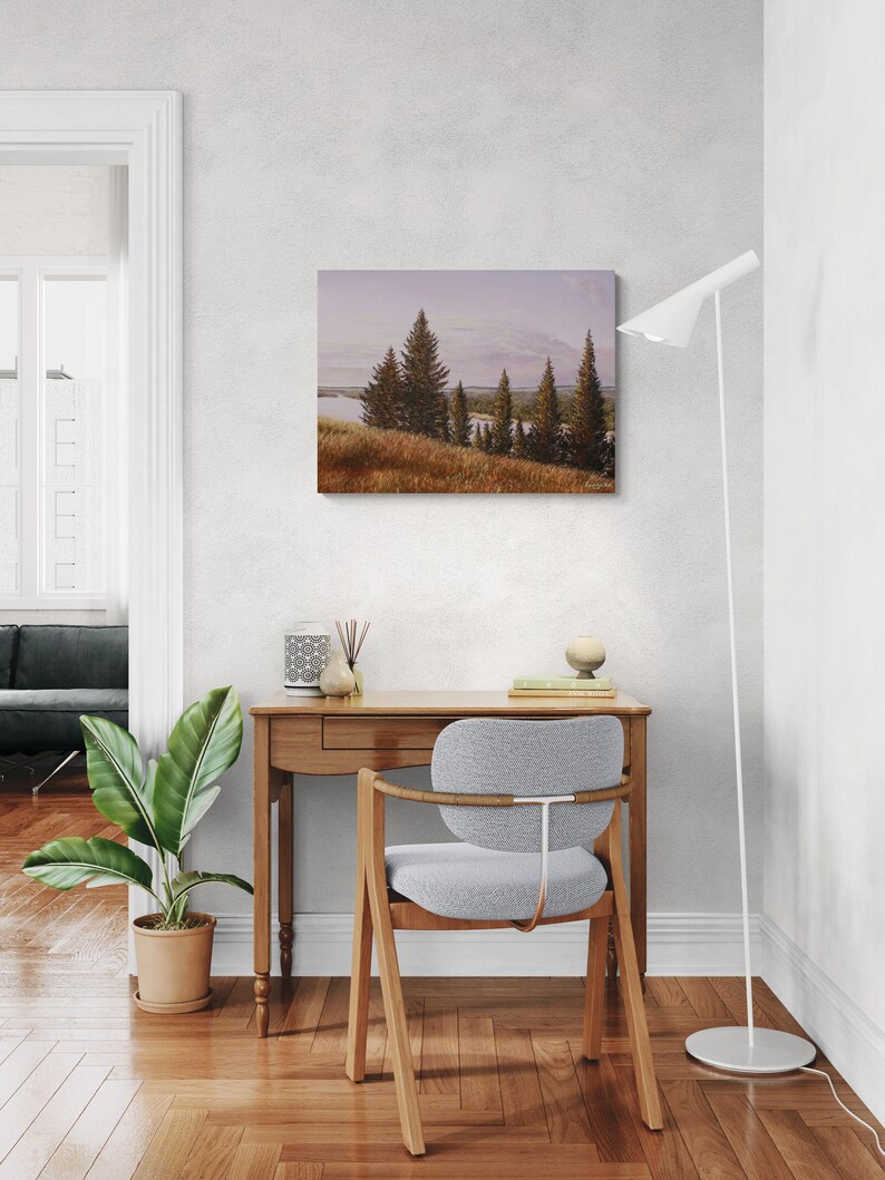 May include: A landscape painting depicts a serene scene with evergreen trees, a body of water, and a golden field under a soft, cloudy sky. The artwork is displayed above a wooden desk with a gray chair, a potted plant, and a modern floor lamp.