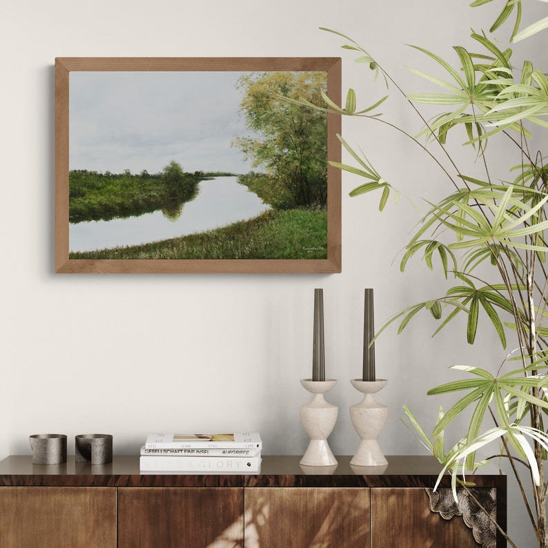 Pu&ograve; includere: Un dipinto di paesaggio incorniciato raffigura una serena scena fluviale. L'opera d'arte presenta una cornice in legno marrone, un cielo nuvoloso e una vegetazione verde lussureggiante lungo le rive del fiume. Il dipinto &egrave; esposto sopra un mobile in legno con oggetti decorativi.