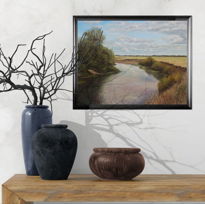 Puede incluir: Una pintura de paisaje enmarcada representa un r&iacute;o que serpentea a trav&eacute;s de un campo bajo un cielo nublado. La pintura se exhibe sobre una superficie de madera, acompa&ntilde;ada de jarrones azules y un cuenco marr&oacute;n oscuro.