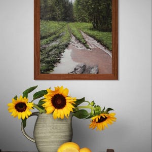 May include: A framed landscape painting of a muddy road through a forest hangs above a wooden table. A vase of sunflowers and lemons sits on the table. The sunflowers have yellow petals and dark brown centers.
