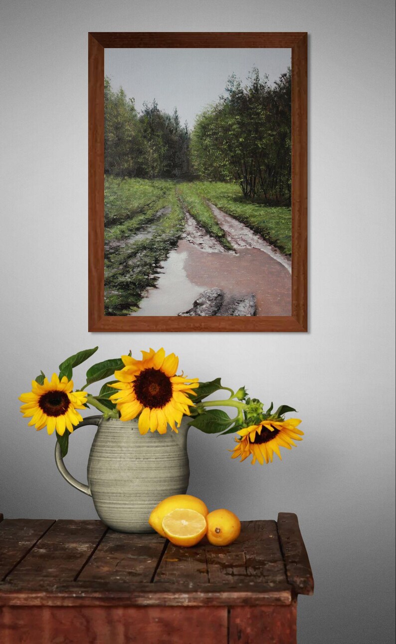 May include: A framed landscape painting of a muddy road through a forest hangs above a wooden table. A vase of sunflowers and lemons sits on the table. The sunflowers have yellow petals and dark brown centers.
