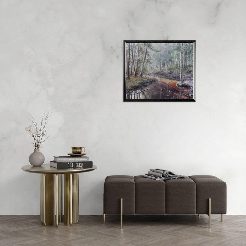 Op de afbeelding: Een ingelijst landschapsschilderij toont een bossc&egrave;ne met een rivier. Het kunstwerk heeft een donkere lijst en wordt tentoongesteld op een witte muur. Een kleine, ronde tafel met een vaas en boeken en een bruine bank met gouden poten staan op de voorgrond.