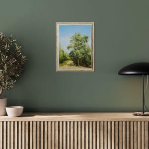 May include: An oil painting of a tree-lined path under a blue sky, framed in a light-colored wooden frame. The artwork is displayed on a dark green wall, next to a potted plant and a black lamp.