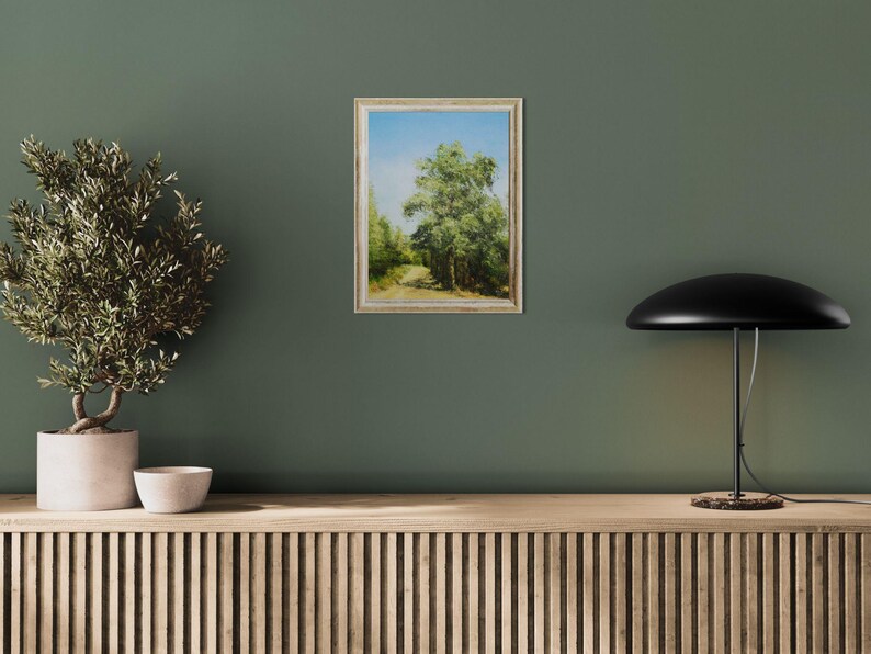 May include: An oil painting of a tree-lined path under a blue sky, framed in a light-colored wooden frame. The artwork is displayed on a dark green wall, next to a potted plant and a black lamp.