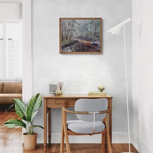 Op de afbeelding: Een ingelijst landschapsschilderij dat een rustige bossc&egrave;ne met een rivier uitbeeldt. Het kunstwerk heeft een bruine houten lijst en wordt aan een witte muur getoond. De kamer omvat een houten bureau, een grijze stoel, een plant in een pot en een witte vloerlamp.