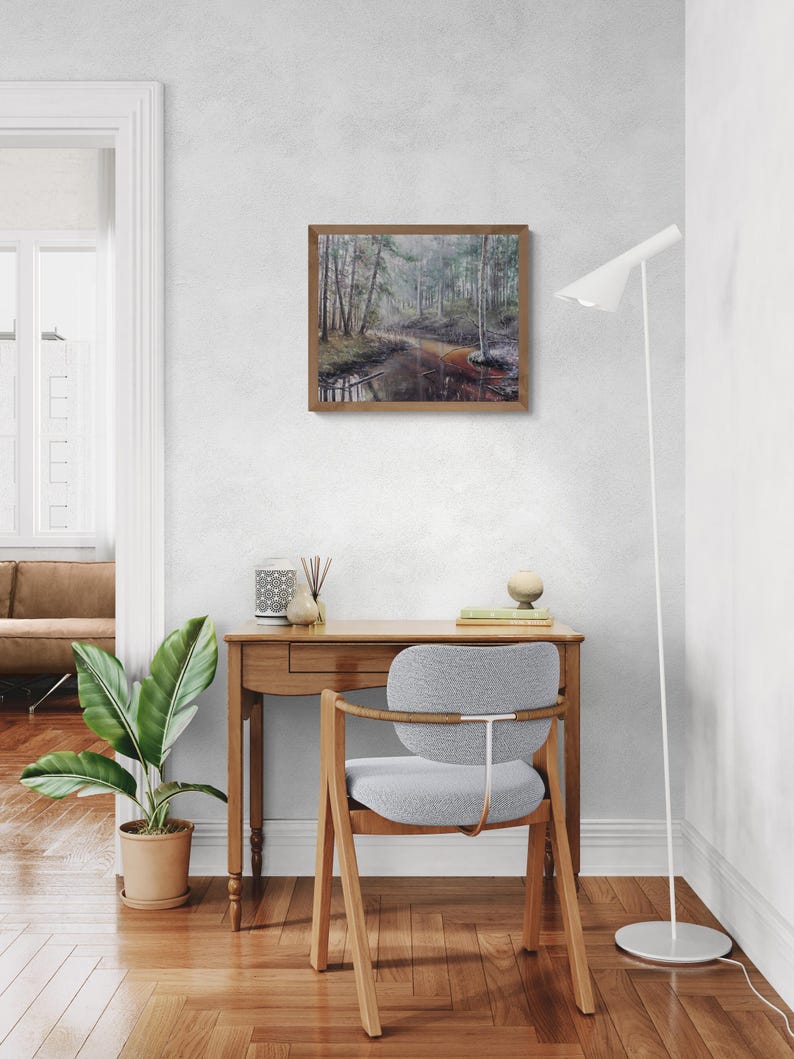 Op de afbeelding: Een ingelijst landschapsschilderij dat een rustige bossc&egrave;ne met een rivier uitbeeldt. Het kunstwerk heeft een bruine houten lijst en wordt aan een witte muur getoond. De kamer omvat een houten bureau, een grijze stoel, een plant in een pot en een witte vloerlamp.