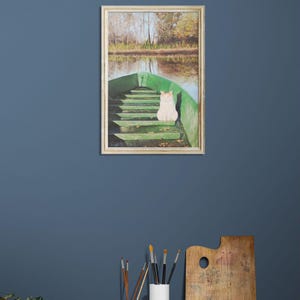 May include: A framed painting depicts a white cat sitting in a green boat on a lake. The boat is in the foreground, with the lake and trees in the background. The painting is displayed on a blue wall above a wooden table.