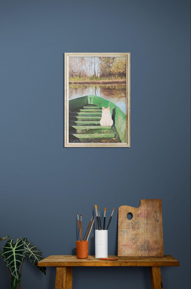 May include: A framed painting depicts a white cat sitting in a green boat on a lake. The boat is in the foreground, with the lake and trees in the background. The painting is displayed on a blue wall above a wooden table.