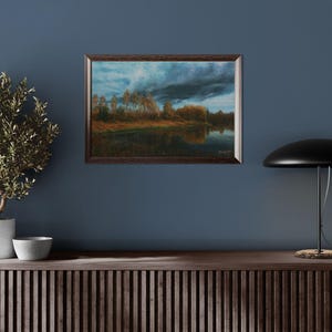 Pu&ograve; includere: Un dipinto paesaggistico incorniciato raffigura una serena scena lacustre sotto un cielo drammatico e nuvoloso. L'opera d'arte presenta una cornice in legno scuro e mostra alberi lungo la riva, con riflessi nell'acqua calma. La tavolozza dei colori comprende blu, marroni e verdi.