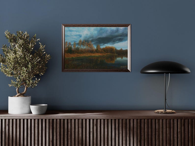 Pu&ograve; includere: Un dipinto paesaggistico incorniciato raffigura una serena scena lacustre sotto un cielo drammatico e nuvoloso. L'opera d'arte presenta una cornice in legno scuro e mostra alberi lungo la riva, con riflessi nell'acqua calma. La tavolozza dei colori comprende blu, marroni e verdi.