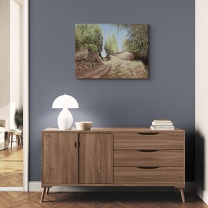 May include: A landscape painting depicting a dirt road winding through a forest, with trees lining both sides. The painting hangs above a wooden cabinet with a white lamp and a bowl. The room has a blue wall and a wooden floor.