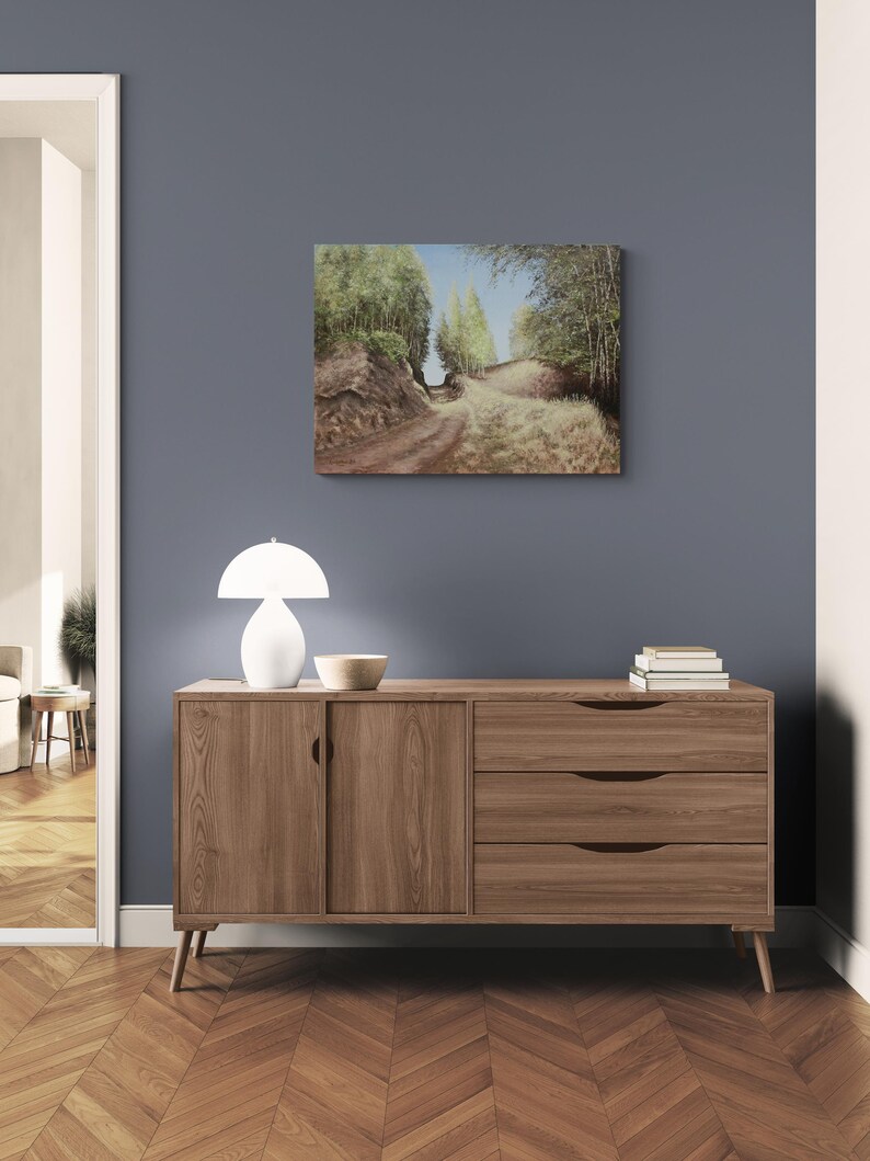 May include: A landscape painting depicting a dirt road winding through a forest, with trees lining both sides. The painting hangs above a wooden cabinet with a white lamp and a bowl. The room has a blue wall and a wooden floor.