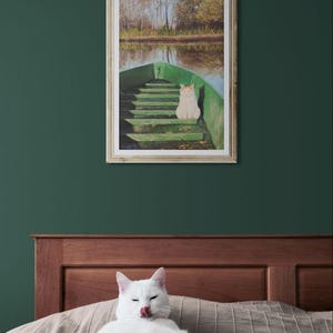 May include: A framed artwork depicts a cat in a green boat on a lake. A white cat is lying on a bed in front of the artwork. The artwork has a wooden frame and is hanging on a dark green wall.