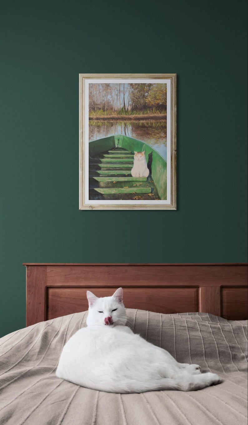 May include: A framed artwork depicts a cat in a green boat on a lake. A white cat is lying on a bed in front of the artwork. The artwork has a wooden frame and is hanging on a dark green wall.